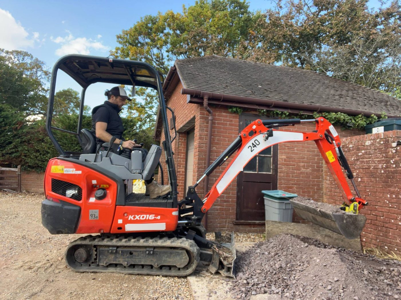 Nick operating a mini digger on a gravel drive beside a brick outbuilding